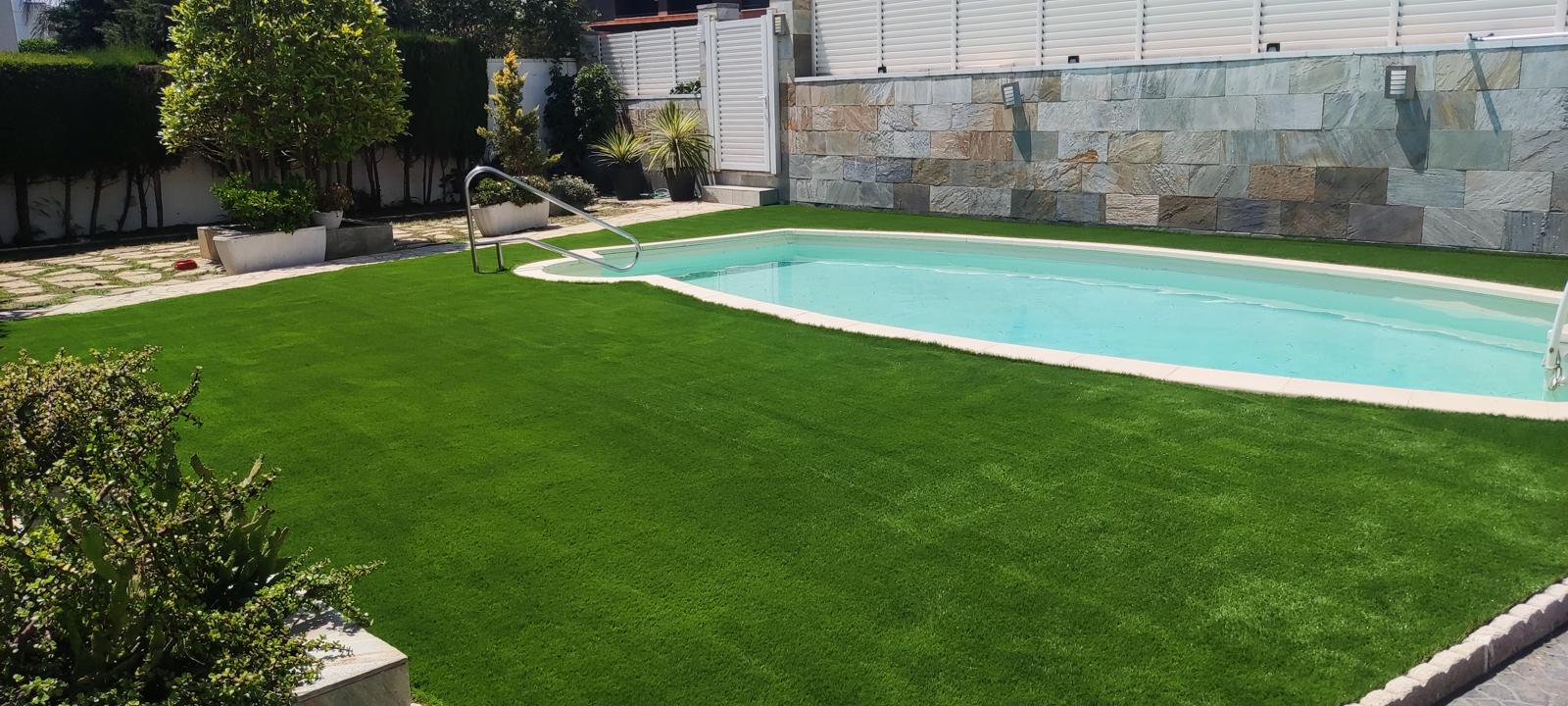 Vista lateral del jardín con césped artificial junto a la piscina Jardín con piscina, tarima y pérgola en Castelldefels Vista lateral del jardín con césped artificial junto a la piscina