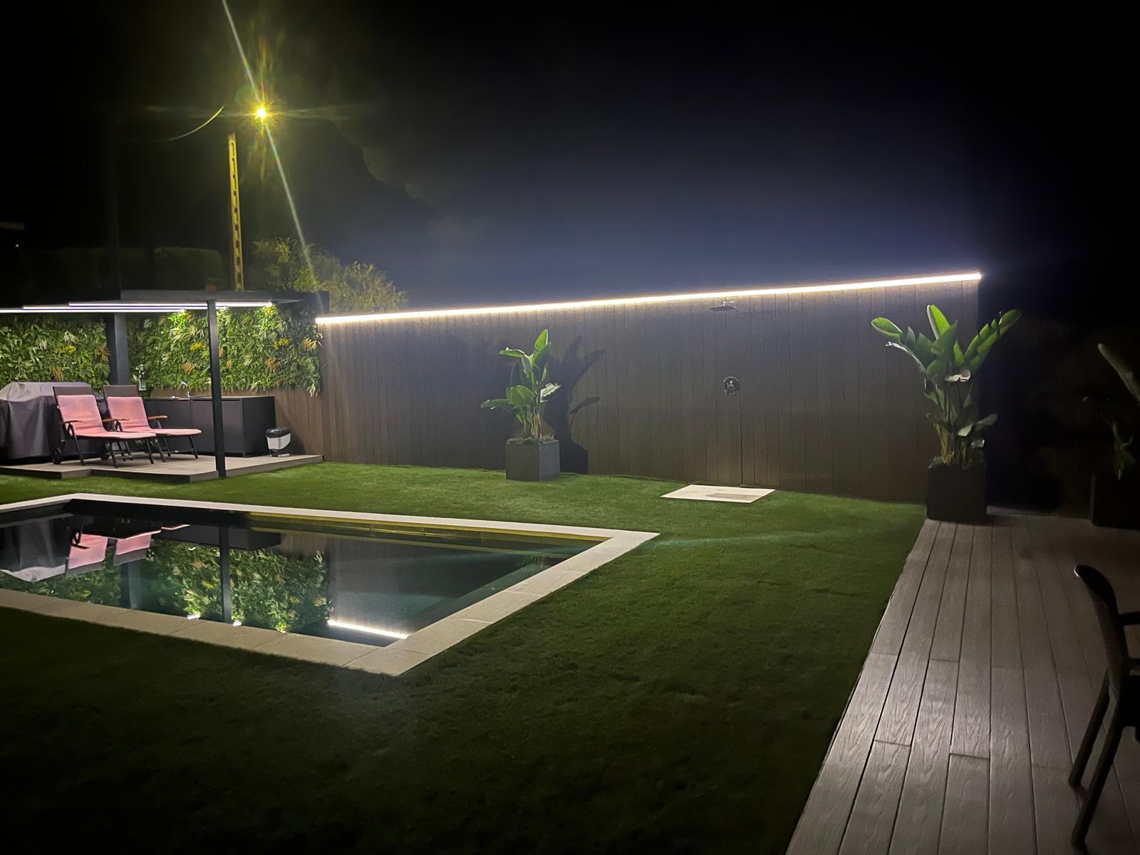 Jardín con piscina y césped artificial en Sitges, pérgola y terraza iluminadas de noche